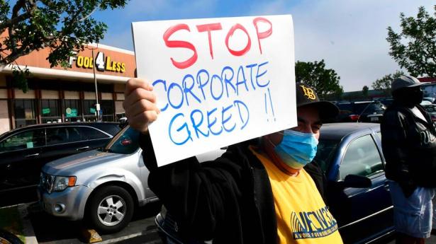 Worker holding up a sign saying stop Corporate Greed in a parking lot in front of a store.