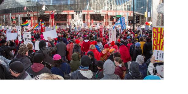 Photo of a massive rally shot from the back with the people facing a building in front and carrying signs. 