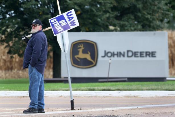 Man in the foreground with a sign that reads UAW On Strike in front of John Deere sign. 