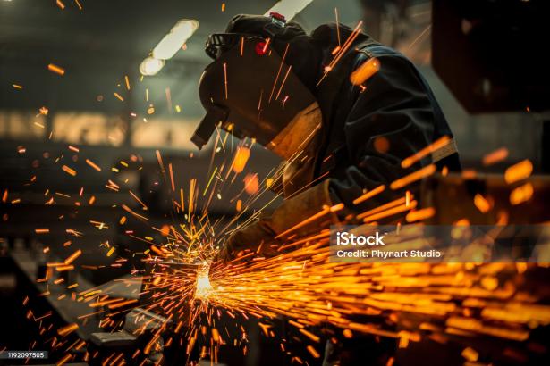 Sparks shooting up from a work table towards a worker with face protection.