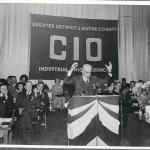 A speaker at a podium with a full house of workers behind him and a huge banner CIO