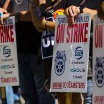 Photo of strike signs on sticks with workers hands leaning on them.   