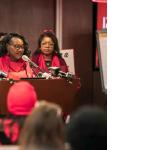 Woman (NYSNA President) at a podium flanked by Nurses in red.