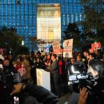 Photo of a rally with signs in front of Conde Nast office building focused on a speaker at the podium.   