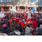 Photo of a massive rally shot from the back with the people facing a building in front and carrying signs. 