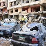 Two cars destroyed by an apparent bomb in from of destroyed buildings.