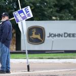 Man in the foreground with a sign that reads UAW On Strike in front of John Deere sign. 