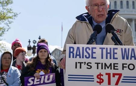 Bernie Sanders at a podium in from of the US Capitol people rallying with signs saying Raise the minimum wage.  Time for $17 and hour.
