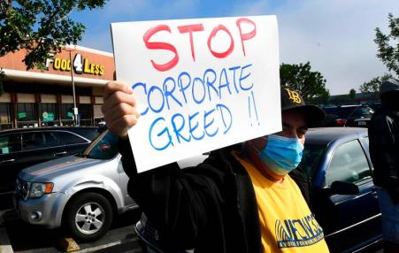 Worker holding up a sign saying stop Corporate Greed in a parking lot in front of a store.