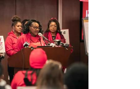 Woman (NYSNA President) at a podium flanked by Nurses in red.