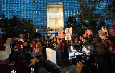 Photo of a rally with signs in front of Conde Nast office building focused on a speaker at the podium.   